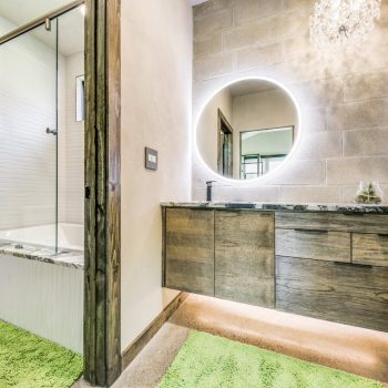 Modern bathroom with wooden vanity, round backlit mirror, green rugs, and a bathtub with glass door.