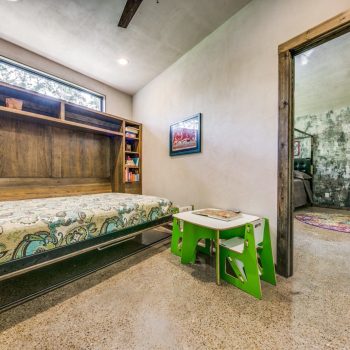 A small room with a pulled-down Murphy bed, wooden shelves, a green kids’ table, and an open doorway to another room.