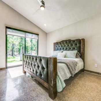 A modern bedroom with a tufted bed, large window, built-in wall shelves, and polished concrete floor.