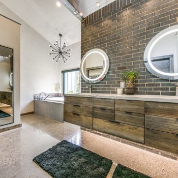 Modern bathroom with dual sinks, round backlit mirrors, wood cabinets, large wall mirror, and a freestanding bathtub.