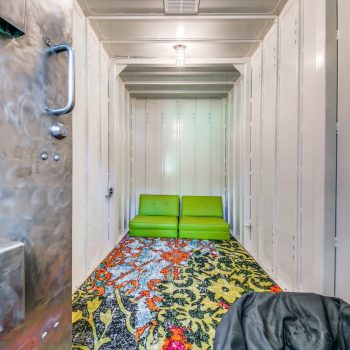 Small room with metallic walls, a green couch, and a colorful patterned carpet, viewed from an open metal door.