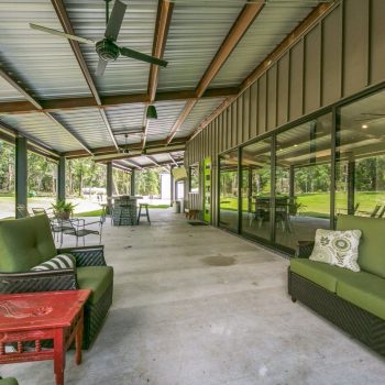 Covered patio with green cushioned seating, ceiling fans, red table, and glass wall overlooking yard and playground.