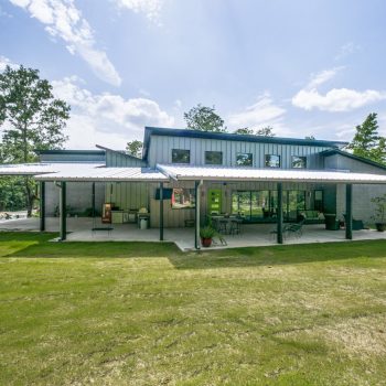 Modern metal building with large covered patio, green lawn, and trees in the background under a partly cloudy sky.