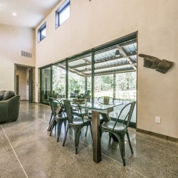 Modern dining area with glass table and metal chairs next to large windows, adjacent to a living room with a sofa.