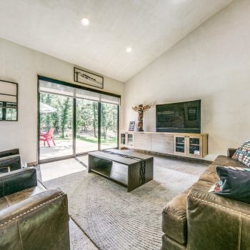 Modern living room with leather chairs, a sofa, coffee table, large TV, and sliding glass doors opening to a backyard.
