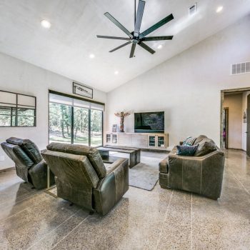 Modern living room with polished concrete floors, three leather chairs, large TV, and ceiling fan, featuring large windows.