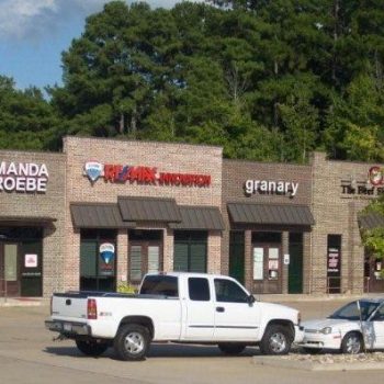 A small strip mall with several storefronts, a white pickup truck, and a white car parked in front.