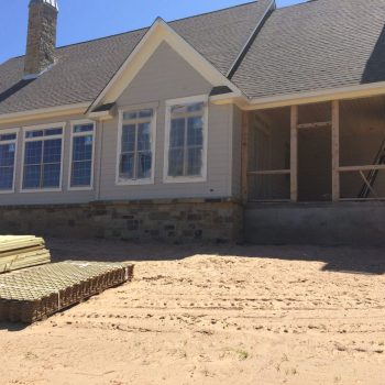 A house under construction with scaffolding on the porch and stacks of lumber on sandy ground in front.
