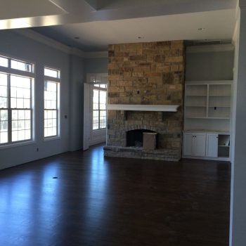 Unfurnished living room with large windows, a stone fireplace, built-in shelves, and dark wood floors.