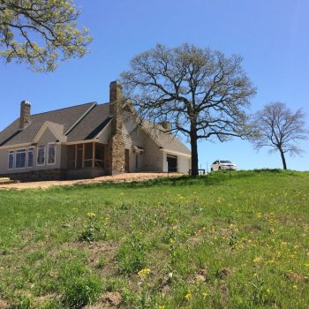 A house with a stone chimney sits on a grassy hill with a few trees under a clear blue sky.