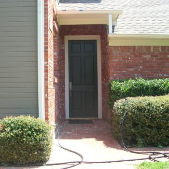 A black door set in a brick house with shrubs and a garden hose along the walkway.