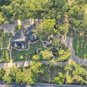 Aerial view of a large property with two houses, a driveway, landscaped gardens, and surrounding trees.