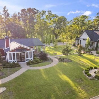 Aerial view of two modern houses with landscaped lawns, curved walkways, and surrounding trees on a sunny day.