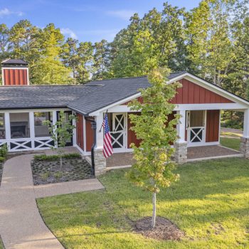Red and white ranch-style house with front porch, American flag, and landscaped yard surrounded by trees.