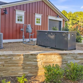Outdoor HVAC unit and generator installed beside a red house with stone retaining wall and surrounding landscaping.
