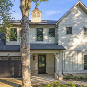 Two-story house with light siding, black garage doors, a front porch, and a large tree in the front yard.