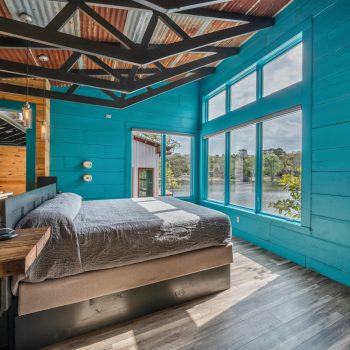 Modern bedroom with teal wooden walls, large windows, a bed, wall-mounted TV, and a view of trees and water outside.
