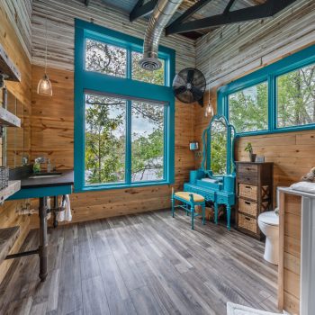 Bathroom with wood panel walls, large windows, teal accents, a vanity, toilet, and open shelving with baskets.