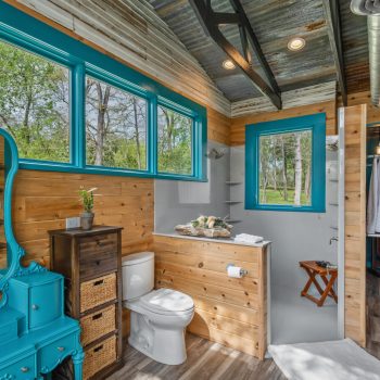 Rustic bathroom with wood paneling, teal accents, vanity, toilet, shower, and clothing area with an outdoor view.