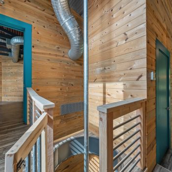 Modern hallway with wood paneling, teal door frames, metal railings, exposed ducts, and spiral staircase opening.