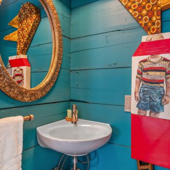 Small bathroom with blue wood walls, round ornate mirror, pedestal sink, and quirky milk carton art with a child painted on it.