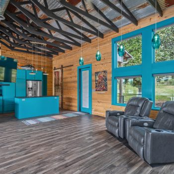 Modern living space with teal kitchen cabinets, wood walls, exposed beams, and two gray recliners facing large windows.