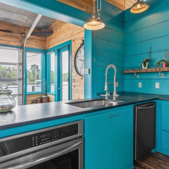Modern kitchen with turquoise cabinets, wood accents, a stainless steel sink, and large windows overlooking water.