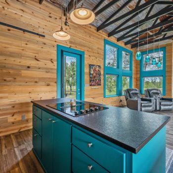 Modern living area with wood walls, teal accents, large windows, and a kitchen island with a cooktop in the foreground.