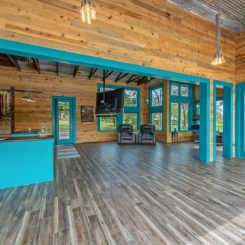 Open-concept living area with wood walls, teal trim, modern kitchen, wood floors, and large windows overlooking trees.