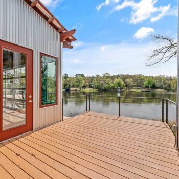 Wooden deck with metal siding overlooks a calm lake surrounded by trees under a partly cloudy sky.