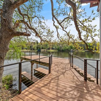 Wooden deck with railing overlooks a calm lake, surrounded by trees, next to a modern glass-walled building.