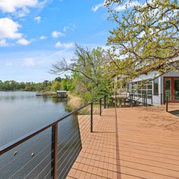 Wooden deck with metal railing overlooks a calm lake, with trees and a building visible under a partly cloudy sky.