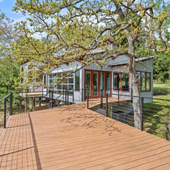 Modern house with large windows and metal siding, featuring a spacious wooden deck surrounded by trees and greenery.