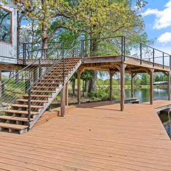 Wooden dock with staircase leading to an elevated deck by a lake, surrounded by trees and a building on the left.