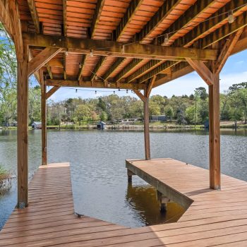 Wooden dock with a covered seating area extends over a calm lake, with houses and trees visible across the water.