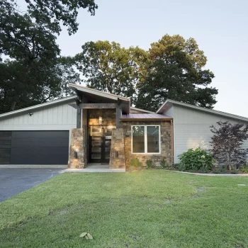 Modern single-story house with stone accents, large front window, and attached garage, surrounded by a grassy lawn and trees.