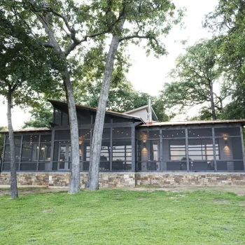 A modern house with screened porch, stone foundation, and large windows, surrounded by grass and tall trees.