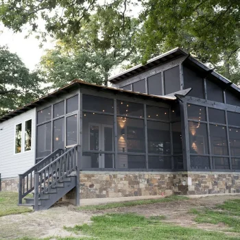 Modern house with large screened-in porch, stone foundation, and surrounded by trees on a grassy lawn.