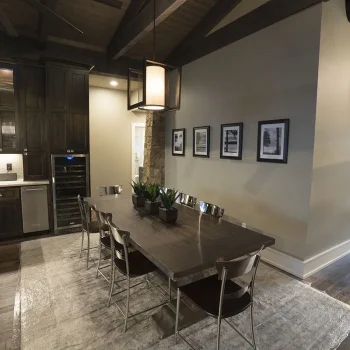 Modern dining area with a wooden table, six chairs, potted plants, and wall art, adjacent to a kitchen and sitting area.