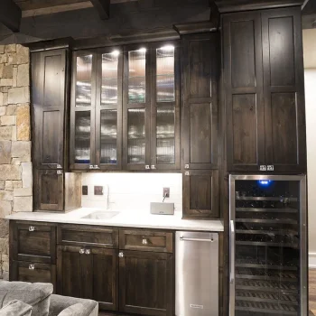 Modern kitchen with dark wood cabinets, glass-front uppers, sink, dishwasher, wine fridge, and stone accent wall.