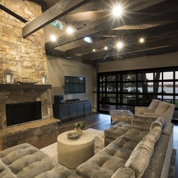Modern living room with stone fireplace, plush seating, wood floors, and large windows overlooking a lake at sunset.