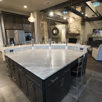 Spacious modern kitchen with a large triangular island, pendant lights, stone fireplace, and a TV in the living area.