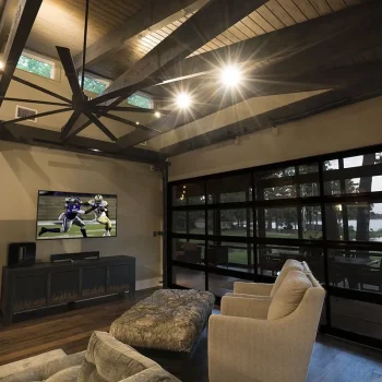 A living room with two chairs, a TV showing football, large windows, and exposed wooden beams on the ceiling.