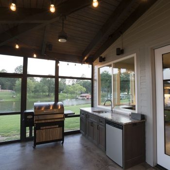 Screened-in outdoor kitchen with grill, sink, and cabinets overlooking a grassy yard and lake, under string lights.