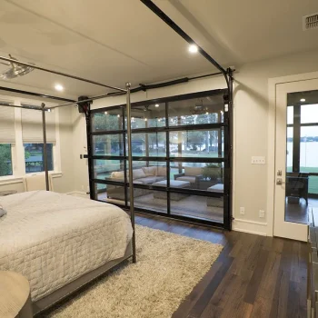 Modern bedroom with a large bed, TV on the wall, and garage-style glass doors opening to a lakeside patio.