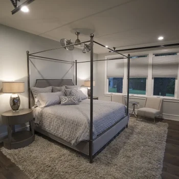 A modern bedroom with a canopy bed, bedside tables, lamps, a shag rug, and large windows with blinds.