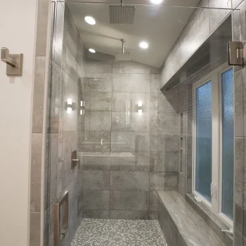 Modern walk-in shower with gray tile walls, pebble floor, glass door, two rainfall showerheads, and a window bench.