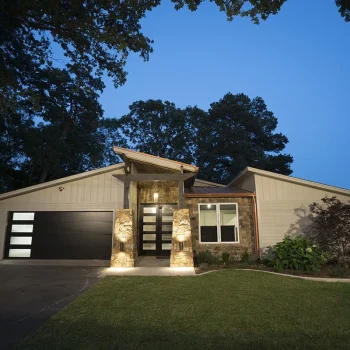 Single-story modern house with stone accents, large windows, and a double garage, photographed at dusk with lights on.