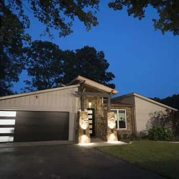 Single-story modern house with a black garage door, illuminated entryway, and surrounding trees at dusk.
