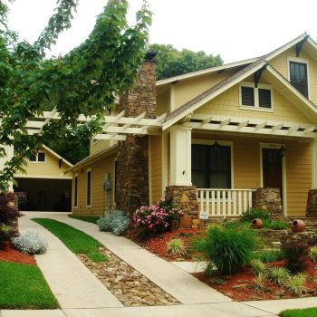 Yellow craftsman-style house with stone chimney, front porch, landscaped yard, and driveway bordered by plants.